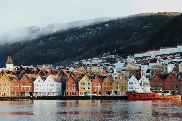 Colorful wooden houses line a waterfront with a red boat moored nearby, at the base of a forested, misty hill.