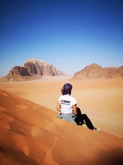 Un capogruppo WeRoad visto di spalle, seduto su una vasta duna di sabbia arancione e che guarda montagne rocciose lontane sotto un cielo azzurro chiaro.