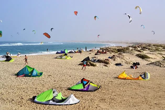Una vasta spiaggia sabbiosa è animata da persone che fanno kitesurf, con molti aquiloni colorati sulla sabbia e nel cielo sopra l'oceano.