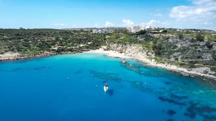 Una vista aerea di una barca a vela ancorata in una baia turchese accanto a una spiaggia sabbiosa appartata, con edifici sulle verdi colline sovrastanti.