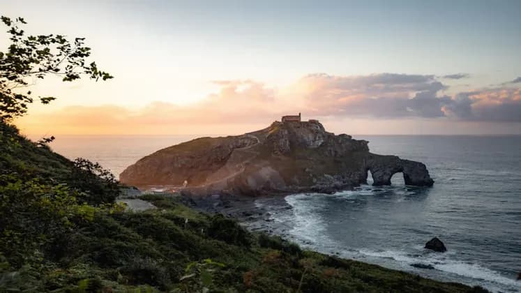 Un isolotto roccioso con un edificio storico in cima, visto da una scogliera verde mentre il sole tramonta sull'oceano.