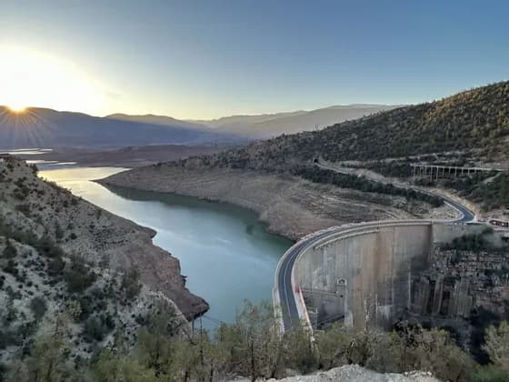 Una grande diga in cemento con una strada in cima si curva attraverso un bacino idrico in una valle montuosa all'alba.