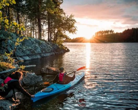 Eine Person an einem felsigen Ufer hilft einer anderen Person in einem blauen Kajak, bei Sonnenuntergang in einen See zu starten.