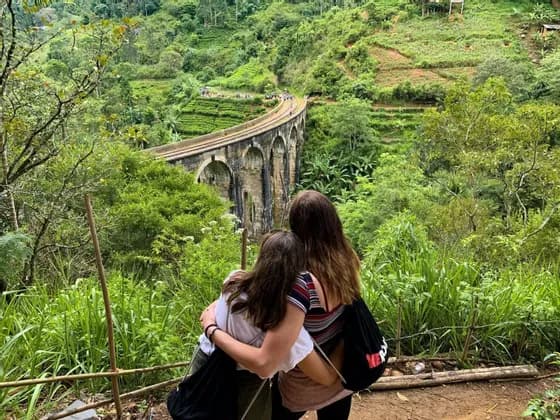 Due donne di un viaggio di gruppo WeRoad si abbracciano mentre ammirano un ponte ad arco in pietra in una valle rigogliosa e verde.
