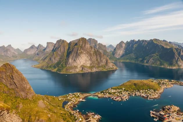 Una vista aerea di un villaggio costiero ai piedi di grandi montagne rocciose circondato da un fiordo blu profondo.
