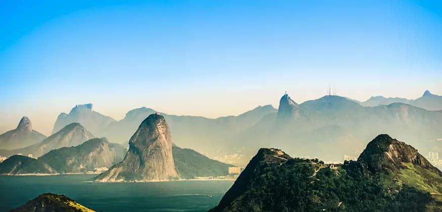 An expansive view of misty mountains rising from the ocean, featuring a distinct conical peak and a distant statue on a summit.