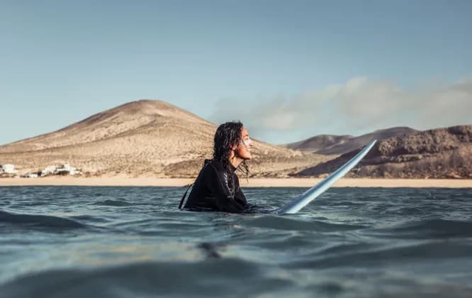 Una persona con i capelli bagnati in una muta nera sorride mentre è seduta su una tavola da surf nell'oceano, con colline sabbiose sullo sfondo.