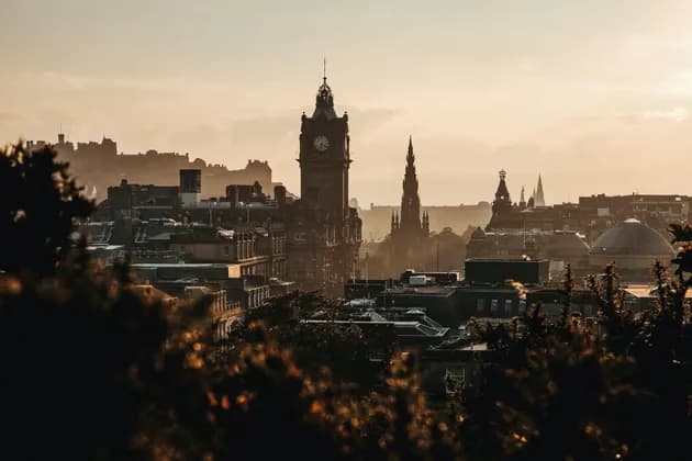 Eine historische Stadtkulisse mit großem Uhrturm und Spitztürmen, die sich bei Sonnenuntergang vor einem warmen, diesigen Himmel abzeichnet.