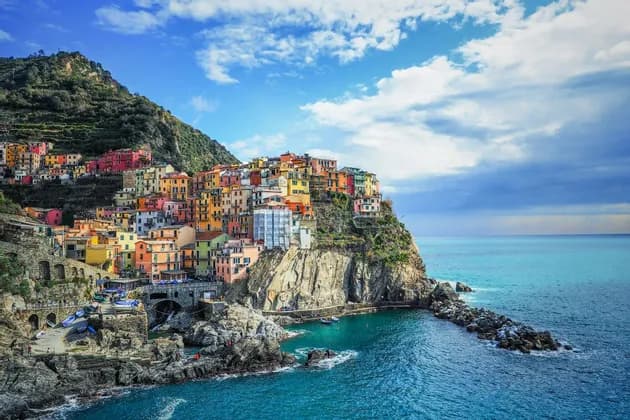 A village of colorful houses built on a rocky cliffside, overlooking a calm blue sea under a partly cloudy sky.