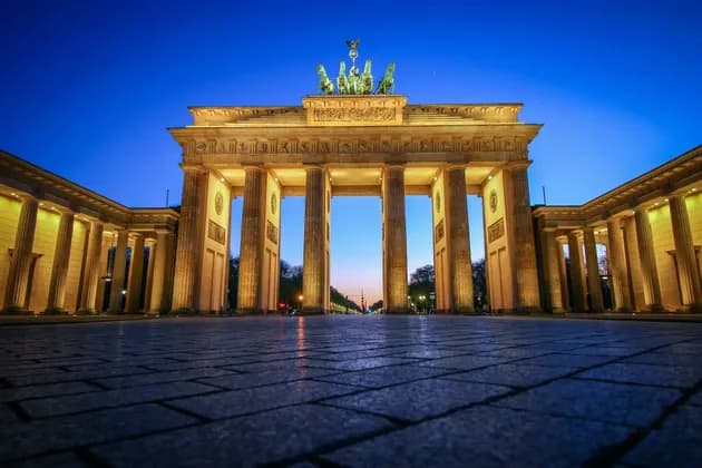 Ein großes, beleuchtetes neoklassizistisches Denkmal mit Säulen in der Dämmerung, in Untersicht von einem gepflasterten Platz aus.