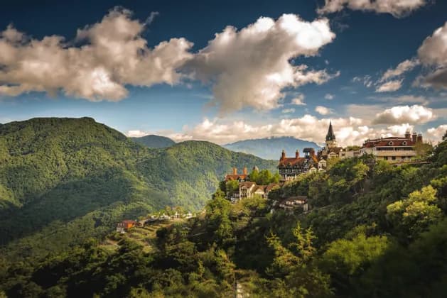 A European-style castle and village nestled in lush green mountains under a partly cloudy sky.