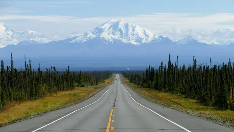 A straight paved road stretches through a forest of evergreens towards a large, snow-capped mountain range in the distance.