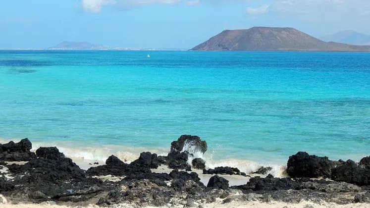 Las olas rompen contra rocas volcánicas negras en una playa de arena, con agua turquesa y una gran isla al fondo.