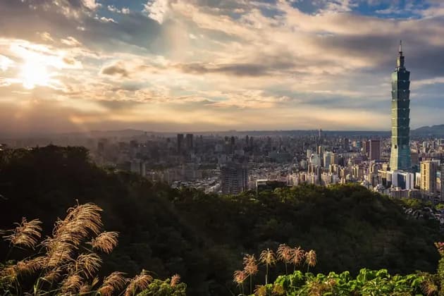 Eine weitläufige Stadtlandschaft mit einem hohen Wolkenkratzer, von einem üppig grünen Hügel bei Sonnenuntergang aus gesehen, mit goldenem Licht, das durch Wolken filtert.