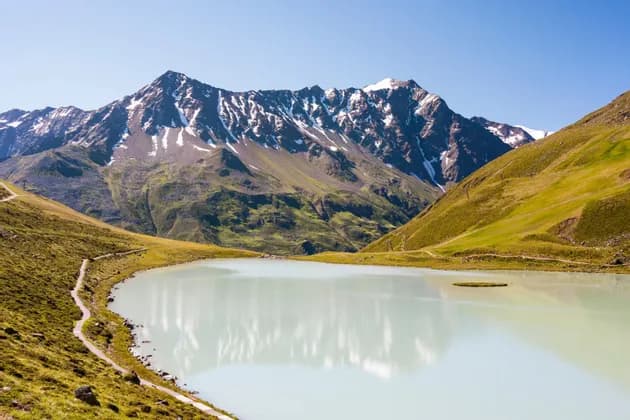 Ein ruhiger, milchiger Alpensee spiegelt an einem sonnigen Tag die schneebedeckten Gipfel einer Bergkette in einem grünen Tal.