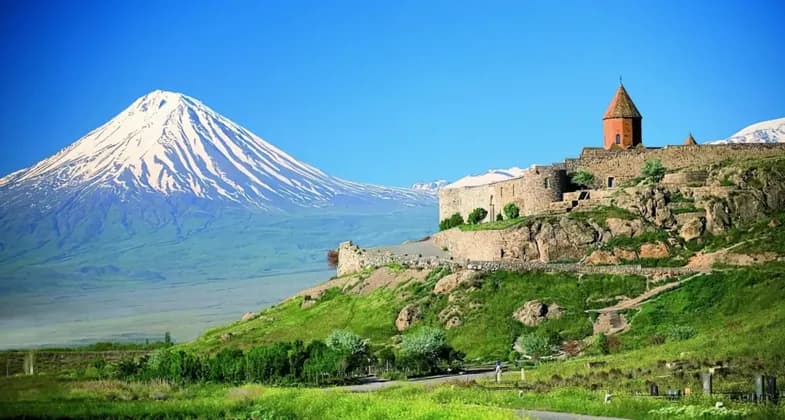 Eine steinerne Festung liegt auf einem grasbewachsenen, felsigen Hügel, mit einem großen, schneebedeckten Berg im Hintergrund vor einem klaren blauen Himmel.