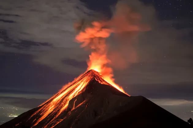 A volcano erupts at night, with glowing lava flowing down its dark slopes under a starry sky.