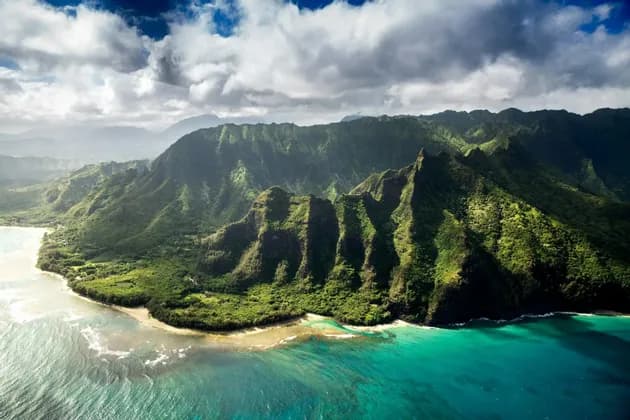 An aerial view of a dramatic, green mountain range descending into a turquoise ocean along a rugged coastline.