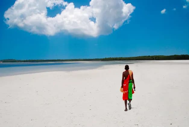 Una persona vista de espaldas con ropa tradicional colorida camina por una extensa playa de arena blanca bajo un cielo azul brillante.