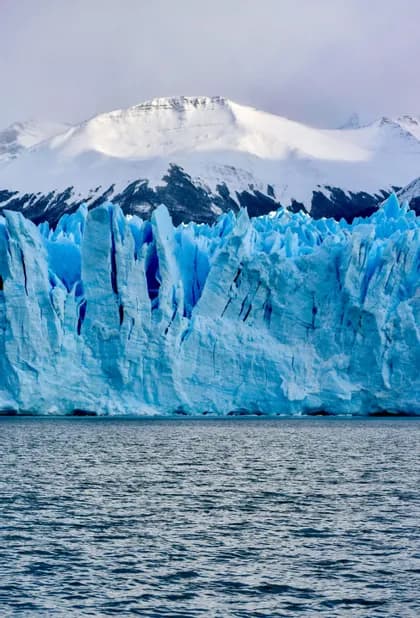 A massive blue glacier rises from the dark water, with snow-covered mountains in the background.