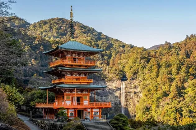 Una pagoda arancione a tre livelli si erge su un fianco di montagna accanto a un'alta cascata, circondata da una fitta foresta verde e gialla.