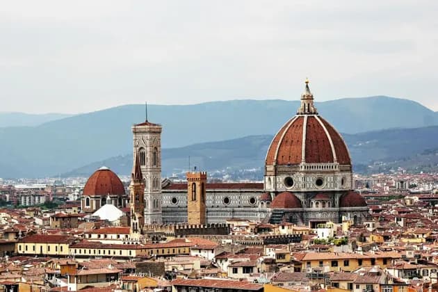 Un paesaggio urbano con tetti in terracotta dominato da una grande cattedrale con una cupola di tegole rosse e un alto campanile, con montagne sullo sfondo.