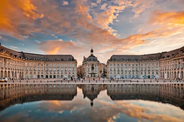 Un grand bâtiment historique se reflète parfaitement dans un miroir d'eau sur une place de ville sous un ciel de coucher de soleil coloré.