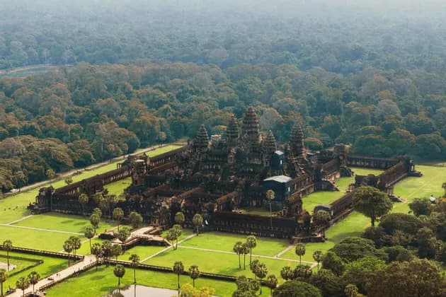 Una vista aerea di un grande e antico complesso di templi in pietra con torri, circondato da prati verdi, palme e una vasta giungla.