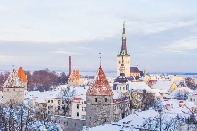 Uno skyline di una città storica in inverno, con tetti coperti di neve, torri di pietra con tetti rossi e un'alta guglia di chiesa.