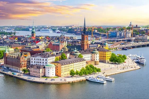 Una vista aerea di una storica città europea con edifici colorati e un campanile, situata su un lungomare con barche ormeggiate al tramonto.