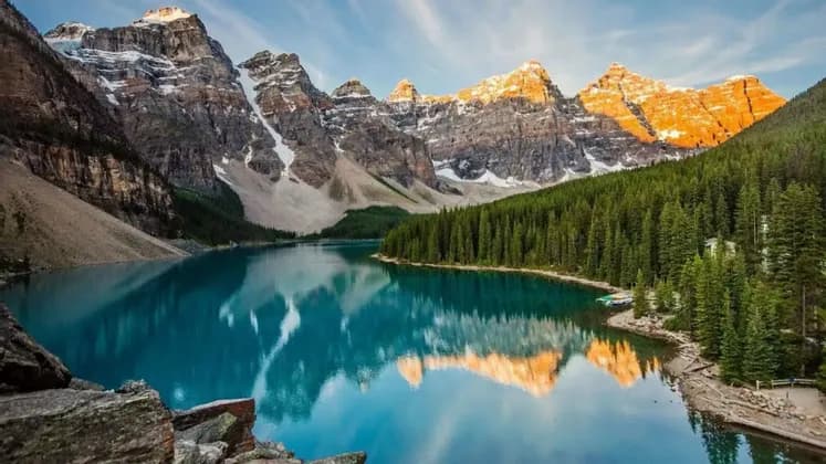 Picos de montañas nevados y soleados que se reflejan en un lago turquesa en calma, rodeado por un denso bosque de pinos.