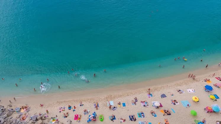Una vista aerea di una spiaggia sabbiosa affollata con persone che nuotano in acqua turchese e prendono il sole su asciugamani colorati.