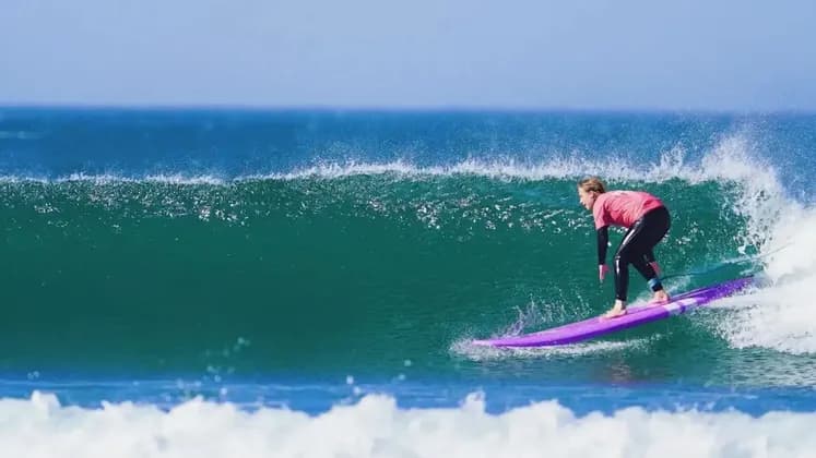 Un surfeur en chemise rose et combinaison chevauche une grande vague turquoise de l'océan sur un longboard violet sous un ciel bleu clair.