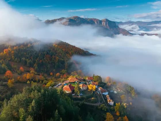Vue aérienne d'un petit village à flanc de montagne, couvert d'arbres aux couleurs automnales, émergeant d'une mer de nuages.