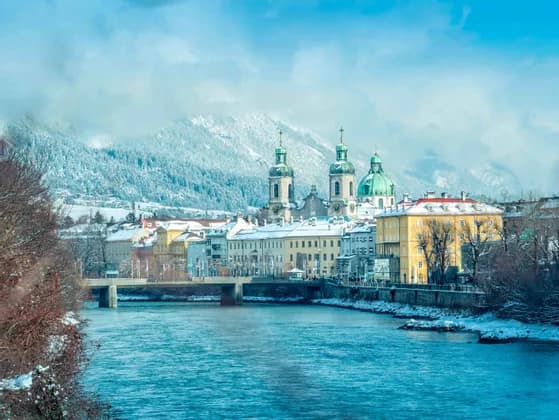 Una vista di una città storica con edifici colorati e cupole di chiese verdi, lungo un fiume, con grandi montagne innevate sullo sfondo.