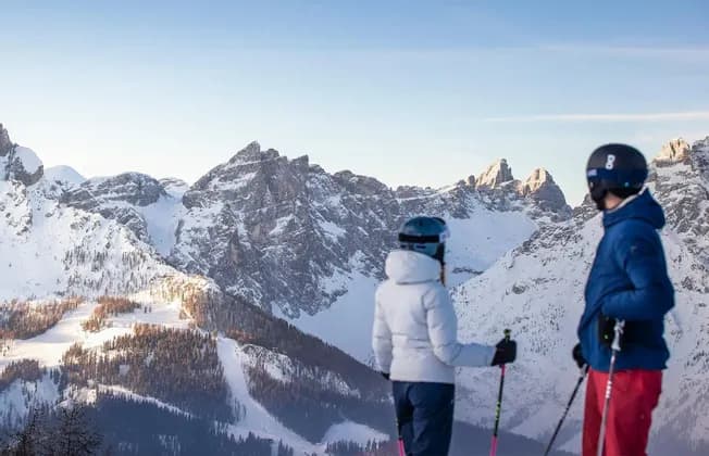 Due sciatori di un viaggio di gruppo WeRoad si fermano ad ammirare una catena montuosa innevata e illuminata dal sole.