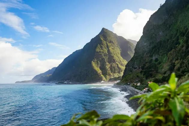 Una carretera costera bordea una costa con olas turquesas, al pie de escarpadas montañas verdes bajo un cielo parcialmente nublado.
