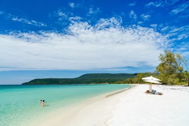Una vasta distesa di spiaggia di sabbia bianca incontra acque limpide e turchesi, con una grande collina verde in lontananza sotto un cielo azzurro.