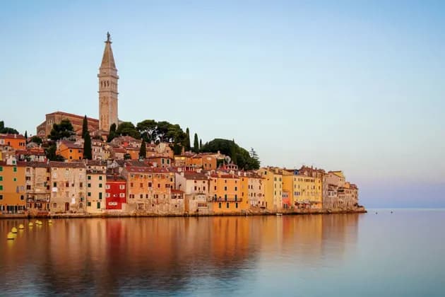 A coastal town with colorful buildings and a prominent bell tower reflecting in the calm water under a clear sky.