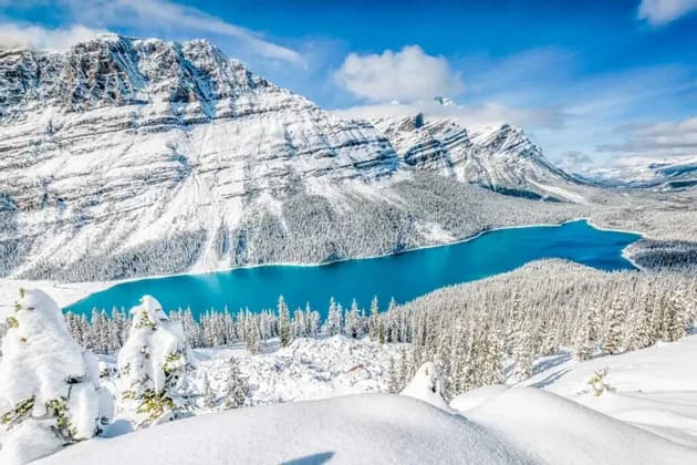 Un vivace lago turchese circondato da una vasta foresta innevata ai piedi di grandi montagne innevate.
