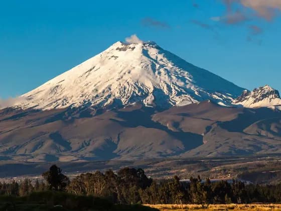 Un gran volcán cubierto de nieve emite una columna de humo desde su cima, dominando un paisaje de colinas y árboles bajo un cielo azul claro.