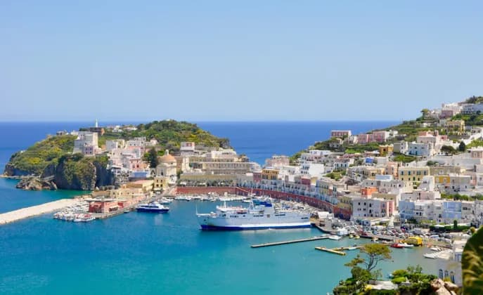 Case colorate su una collina circondano un porto blu brillante dove un grande traghetto e barche più piccole sono ormeggiate sotto un cielo sereno.