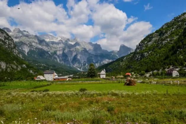 A rural village with a church sits in a lush green valley at the base of jagged, rocky mountains under a partly cloudy sky.