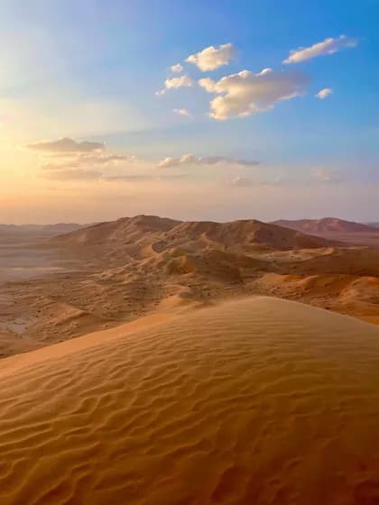 Ampia vista di dune di sabbia nel deserto al tramonto, sotto un cielo azzurro con nuvole bianche.