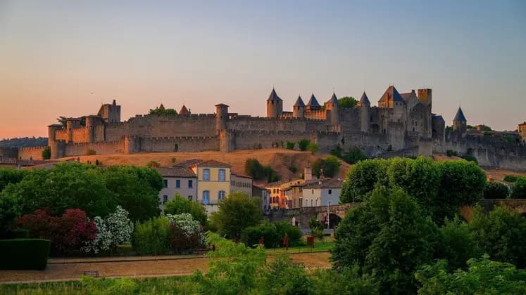 Un grande castello in pietra con molte torrette si erge su una collina, illuminato dalla calda luce del tramonto sopra una piccola città con alberi verdi e lussureggianti.