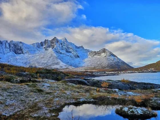 Vista panorámica de montañas nevadas elevándose sobre un pueblo costero y un fiordo, con un primer plano rocoso y parcialmente nevado.
