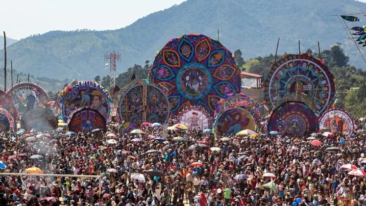 Eine riesige Menschenmenge versammelt sich auf einem Open-Air-Festival, um mehrere riesige, bunte, verzierte Drachen zu bestaunen, die vor einer Bergkette ausgestellt sind.