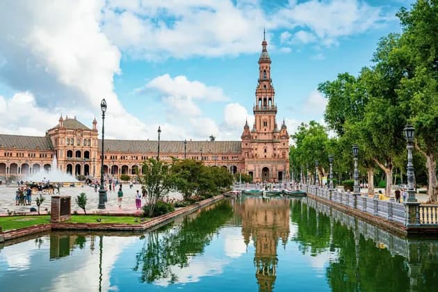 Ein großes, verziertes Gebäude mit einem hohen Turm spiegelt sich in einem Kanal auf einem belebten Platz unter einem bewölkten Himmel.