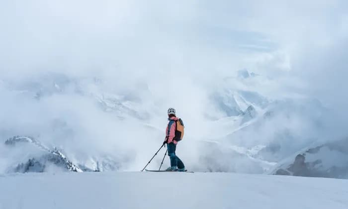 Una persona con gli sci, casco e zaino, si trova su un crinale innevato, ammirando una vasta catena montuosa avvolta dalle nuvole.