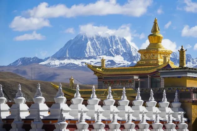A row of white stupas in front of a golden-roofed temple with a large, snow-covered mountain in the background.